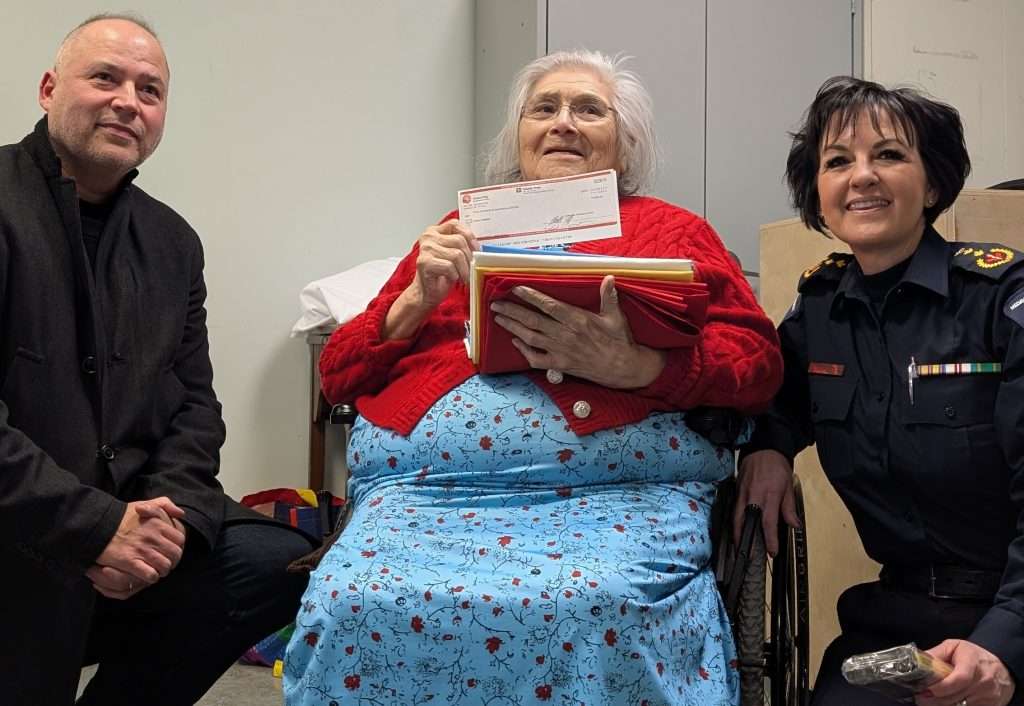 An Indigenous man and a woman in uniform are on either side of a kokum in a wheelchair holding a cheque and cloth. They are all smiling.